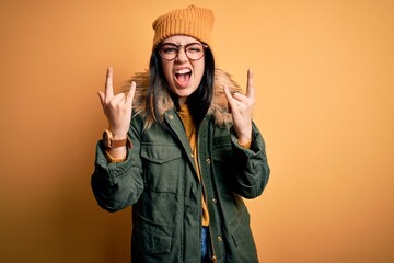 Young brunette woman wearing glasses and winter coat with hat over yellow isolated background shouting with crazy expression doing rock symbol with hands up. Music star. Heavy concept.