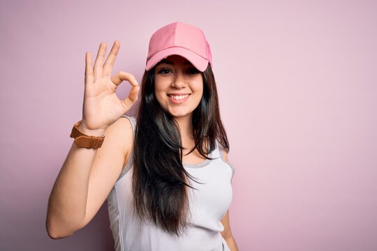 Young brunette woman wearing casual sport cap over pink background smiling positive doing ok sign with hand and fingers. Successful expression.