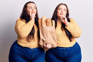 Young plus size twins holding paper bag with bread serious face thinking about question with hand on chin, thoughtful about confusing idea