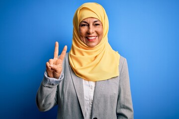 Middle age brunette business woman wearing muslim traditional hijab over blue background smiling looking to the camera showing fingers doing victory sign. Number two.