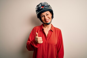 Middle age motorcyclist woman wearing motorcycle helmet over isolated white background doing happy thumbs up gesture with hand. Approving expression looking at the camera showing success. © Krakenimages.com