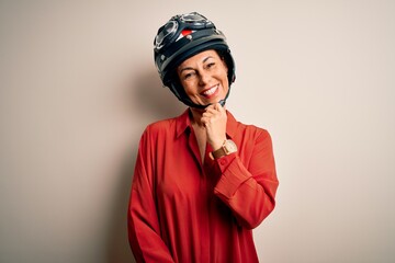 Middle age motorcyclist woman wearing motorcycle helmet over isolated white background looking confident at the camera with smile with crossed arms and hand raised on chin. Thinking positive.