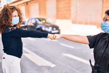 Two beautiful friends wearing medical mask. Bump fists standing at town street.