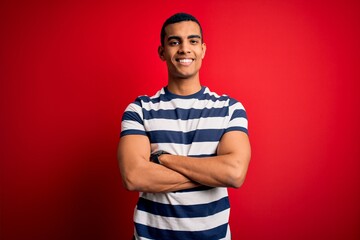 Handsome african american man wearing casual striped t-shirt standing over red background happy face smiling with crossed arms looking at the camera. Positive person.