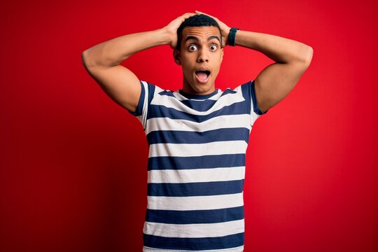Handsome african american man wearing casual striped t-shirt standing over red background Crazy and scared with hands on head, afraid and surprised of shock with open mouth