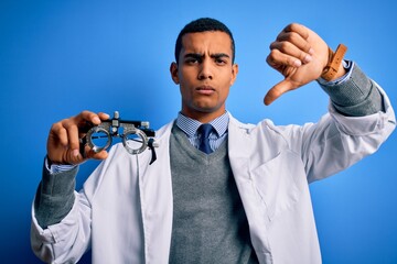 Handsome african american optical man holding optometry glasses over blue background with angry face, negative sign showing dislike with thumbs down, rejection concept