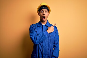 Young handsome african american worker man wearing blue uniform and security helmet Surprised pointing with finger to the side, open mouth amazed expression. - obrazy, fototapety, plakaty