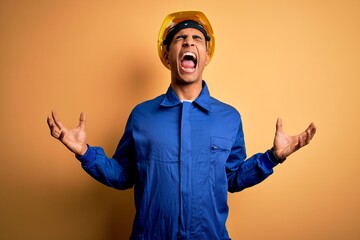 Young handsome african american worker man wearing blue uniform and security helmet crazy and mad...