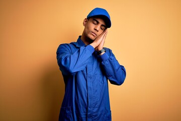 Young african american mechanic man wearing blue uniform and cap over yellow background sleeping tired dreaming and posing with hands together while smiling with closed eyes.