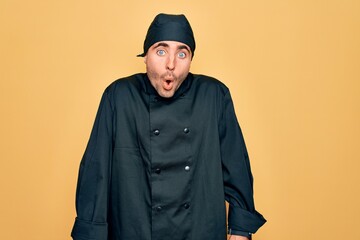 Young handsome cooker man with blue eyes wearing uniform and hat over yellow background afraid and shocked with surprise expression, fear and excited face.