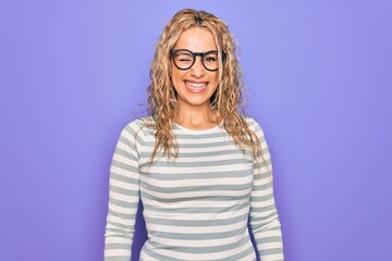 Beautiful blonde woman wearing casual striped t-shirt and glasses over purple background winking...
