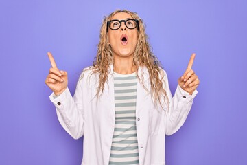 Young beautiful blonde scientist woman wearing coat and glasses over purple background amazed and surprised looking up and pointing with fingers and raised arms.