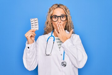 Young beautiful blonde cardiologist woman wearing stethoscope holding plastic heart covering mouth...