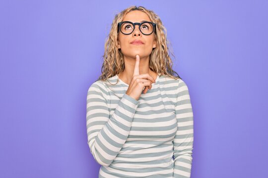 Beautiful blonde woman wearing casual striped t-shirt and glasses over purple background Thinking concentrated about doubt with finger on chin and looking up wondering