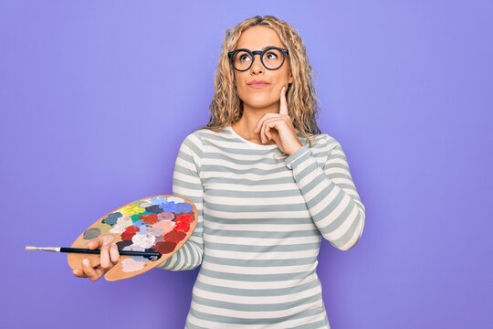 Young beautiful blonde artist woman drawing using paintbrush and palette with colors serious face thinking about question with hand on chin, thoughtful about confusing idea