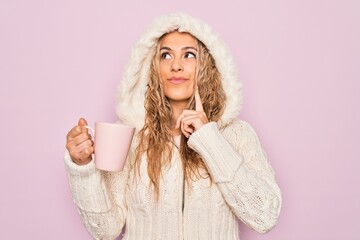 Young beautiful blonde woman wearing casual sweater with hood drinking cup of coffee serious face thinking about question with hand on chin, thoughtful about confusing idea