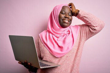 African american plus size woman wearing muslim hijab using laptop over pink background smiling...