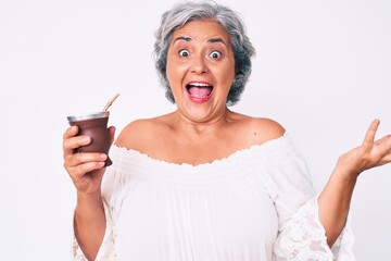 Senior hispanic woman drinking mate infusion celebrating achievement with happy smile and winner expression with raised hand