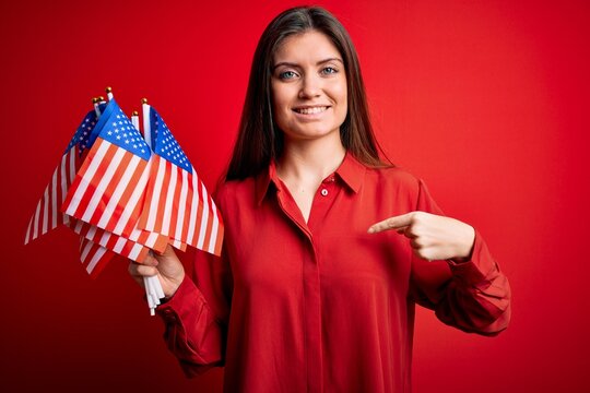 Young beautiful patriotic woman with blue eyes holding united states flags over red background with surprise face pointing finger to himself