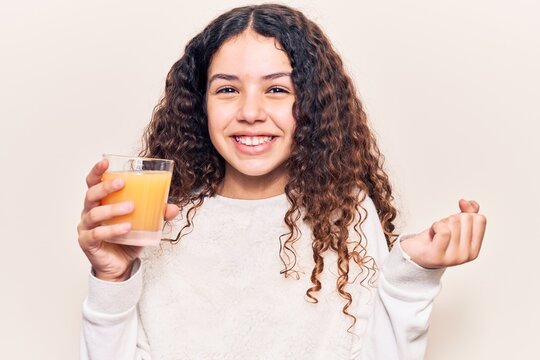 Beautiful kid girl with curly hair drinking glass of orange juice screaming proud, celebrating victory and success very excited with raised arm