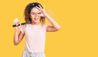 Beautiful kid girl with curly hair holding badminton racket and shuttlecock smiling happy doing ok sign with hand on eye looking through fingers