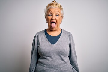 Senior beautiful grey-haired woman wearing golden queen crown over white background sticking tongue out happy with funny expression. Emotion concept.