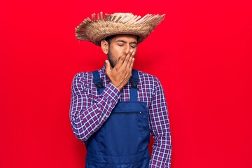 Young latin man wearing farmer hat and apron bored yawning tired covering mouth with hand. restless...