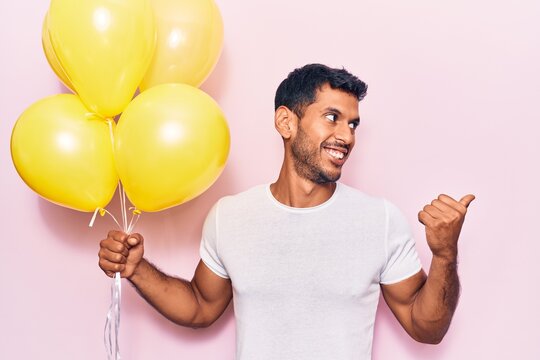 Young latin man holding balloons pointing thumb up to the side smiling happy with open mouth