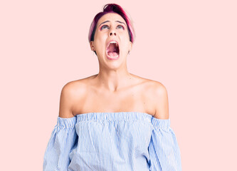 Young beautiful woman with pink hair wearing casual clothes angry and mad screaming frustrated and...
