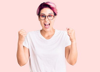 Young beautiful woman with pink hair wearing casual clothes and glasses celebrating surprised and...