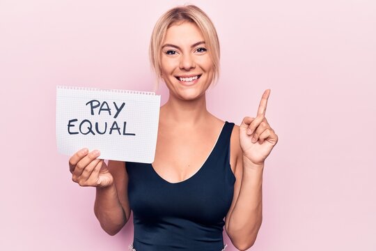Beautiful blonde woman asking for equality economy holding paper with pay equal message smiling with an idea or question pointing finger with happy face, number one