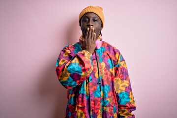 Young handsome african american man wearing colorful coat and cap over pink background bored yawning tired covering mouth with hand. Restless and sleepiness.