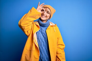 Young blonde woman with short hair wearing rain coat for rainy weather over blue background doing ok gesture with hand smiling, eye looking through fingers with happy face.