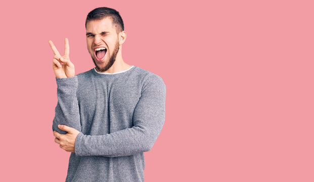 Young handsome man wearing casual sweater smiling with happy face winking at the camera doing victory sign. number two.