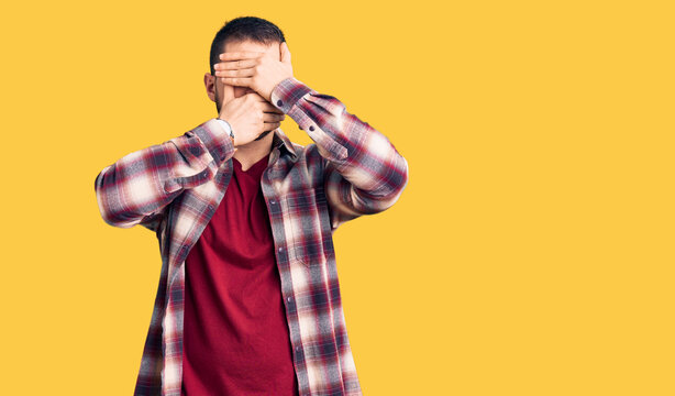Young handsome man wearing casual shirt covering eyes and mouth with hands, surprised and shocked. hiding emotion