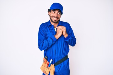 Handsome young man with curly hair and bear weaing handyman uniform laughing nervous and excited with hands on chin looking to the side