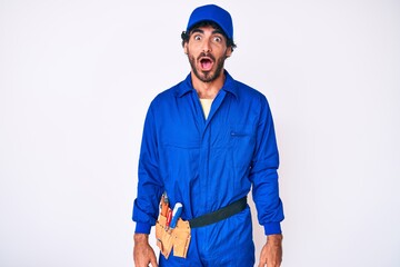 Handsome young man with curly hair and bear weaing handyman uniform afraid and shocked with surprise and amazed expression, fear and excited face.