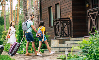 Obraz na płótnie Canvas Nice hotel. A photo of a happy family with luggages and backpacs on a weekend coming to the wooden hotel surrounded by trees, father holding his daughter's hand.