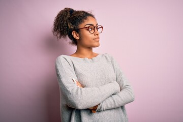 Young beautiful african american girl wearing sweater and glasses over pink background looking to the side with arms crossed convinced and confident