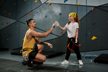 Male instructor teaching little girl, giving high five while applying white dust of magnesia, chalk on hands before climbing wall in bouldering center