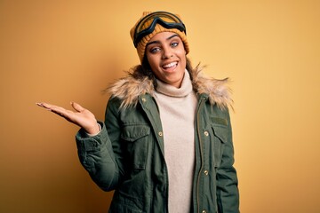 Young african american afro skier girl wearing snow sportswear and ski goggles smiling cheerful presenting and pointing with palm of hand looking at the camera.