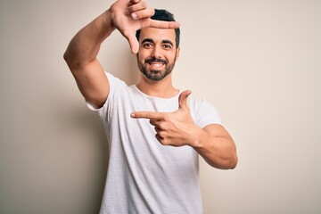 Young handsome man with beard wearing casual t-shirt standing over white background smiling making frame with hands and fingers with happy face. Creativity and photography concept.