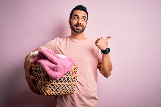 Young handsome man with beard doing housework holding wicker basket with clothes pointing and showing with thumb up to the side with happy face smiling