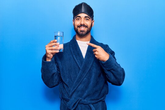 Young hispanic man wearing sleep mask and robe drinking water smiling happy pointing with hand and finger