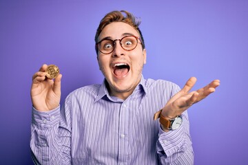 Young handsome redhead businessman holding cryptocurrency bitcoin over purple background celebrating victory with happy smile and winner expression with raised hands