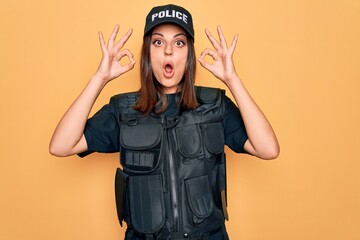 Young beautiful brunette policewoman wearing police uniform bulletproof and cap looking surprised and shocked doing ok approval symbol with fingers. Crazy expression