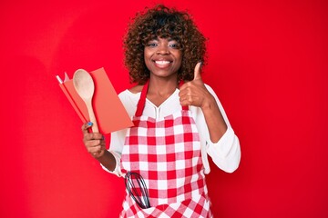 Young african american woman wearing professional baker apron reading cooking recipe book smiling...
