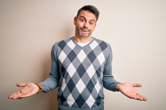 Young handsome man wearing casual sweater standing over isolated white background clueless and confused expression with arms and hands raised. Doubt concept.