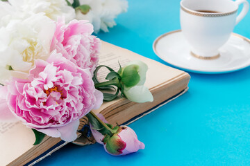 Summer romantic breakfast. Open book with white and pink peonies, white cup of coffee on a blue background.Good morning concept.Copy space, selective focus with shallow depth of field