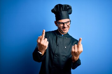 Young handsome chef man wearing cooker uniform and hat over isolated blue background Showing middle finger doing fuck you bad expression, provocation and rude attitude. Screaming excited